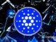 Logo oficial do Cardano (ADA) em azul sobre fundo com elementos gráficos digitais e fragmentos geométricos.